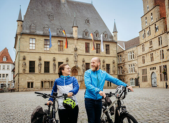Friedensroute: Historisches Rathaus in Osnabrück mit zwei Radfahrenden, die ihre Fahrräder schieben, davor. . 