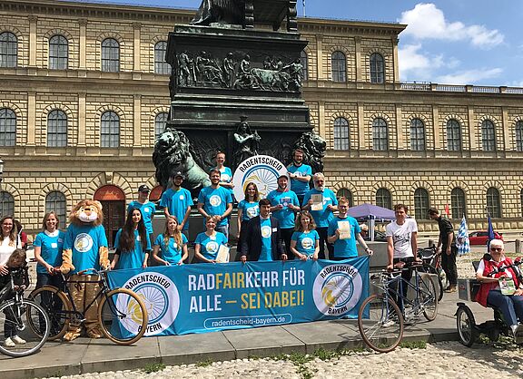 Etwa 15 Personen in blauen Aktions-T-Shirts präsentieren auf einem Platz in München das Transparent des Radentscheids Bayern. 