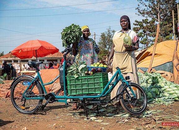Zwei afrikanische Frauen stehen auf einem Markt im Freien hinter einem türkisgrünen Lastenfahrrad. Auf der tiefen Ladefläche zwischen Lenker und Vorderrad steht eine grüne Kiste, die voll mit Auberginen, Orangen und Blattgemüse beladen ist. Die Frauen halten weiteres Gemüse in den Händen und blicken freundlich in die Kamera.