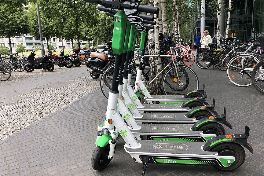 E-Scooter zum Ausleihen in Berlin Im Vordergrund mehrere E-Scooter eines Verleihs, im Hintergrund mehrere Fahrräder, einige Personen und Gebäude