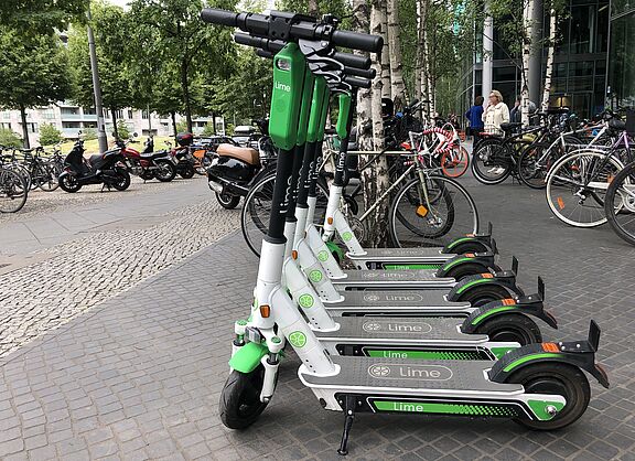 Im Vordergrund mehrere E-Scooter eines Verleihs, im Hintergrund mehrere Fahrräder, einige Personen und Gebäude