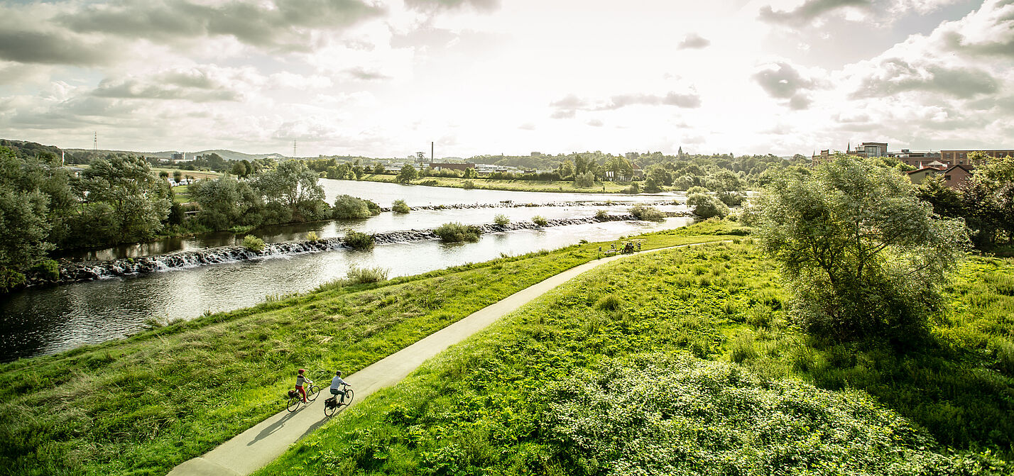 Der RuhrtalRadweg in NRW ist eine von 46 ADFC-Qualitätsradrouten in Deutschland. Panoramablick aus der Vogelperspektive auf den Ruhrtalradweg entlang der Ruhr, zwei Radfahrende fahren der Nachmittagssonne entgegen.